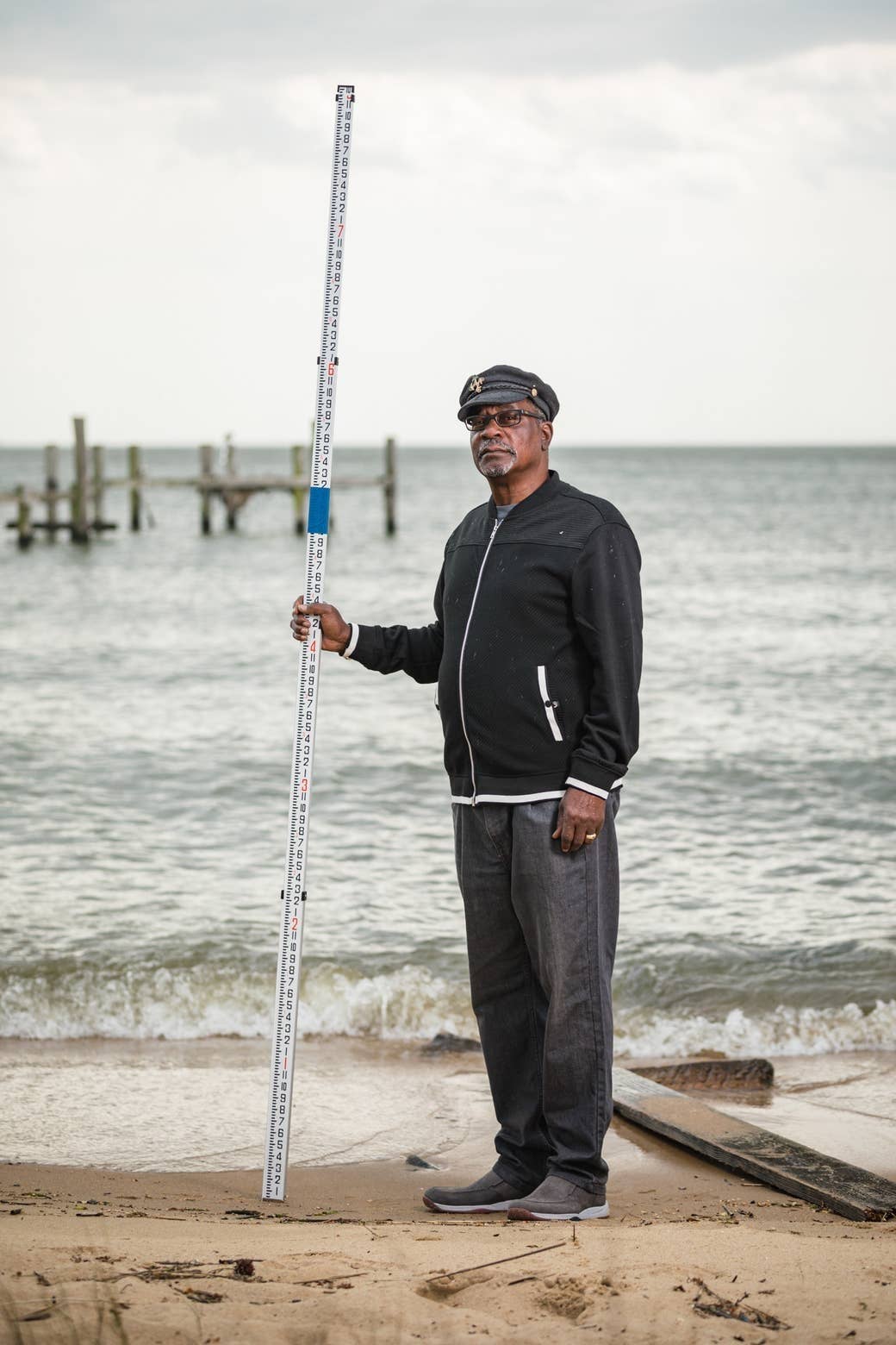 A man standing at the beach holding a long measuring stick with a taped marking at his shoulders