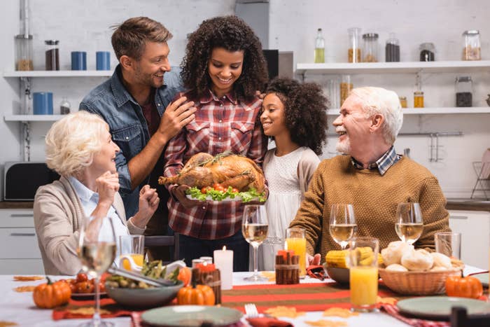 Family gathered around a Thanksgiving table while mother holds a beautifully-roasted turkey.