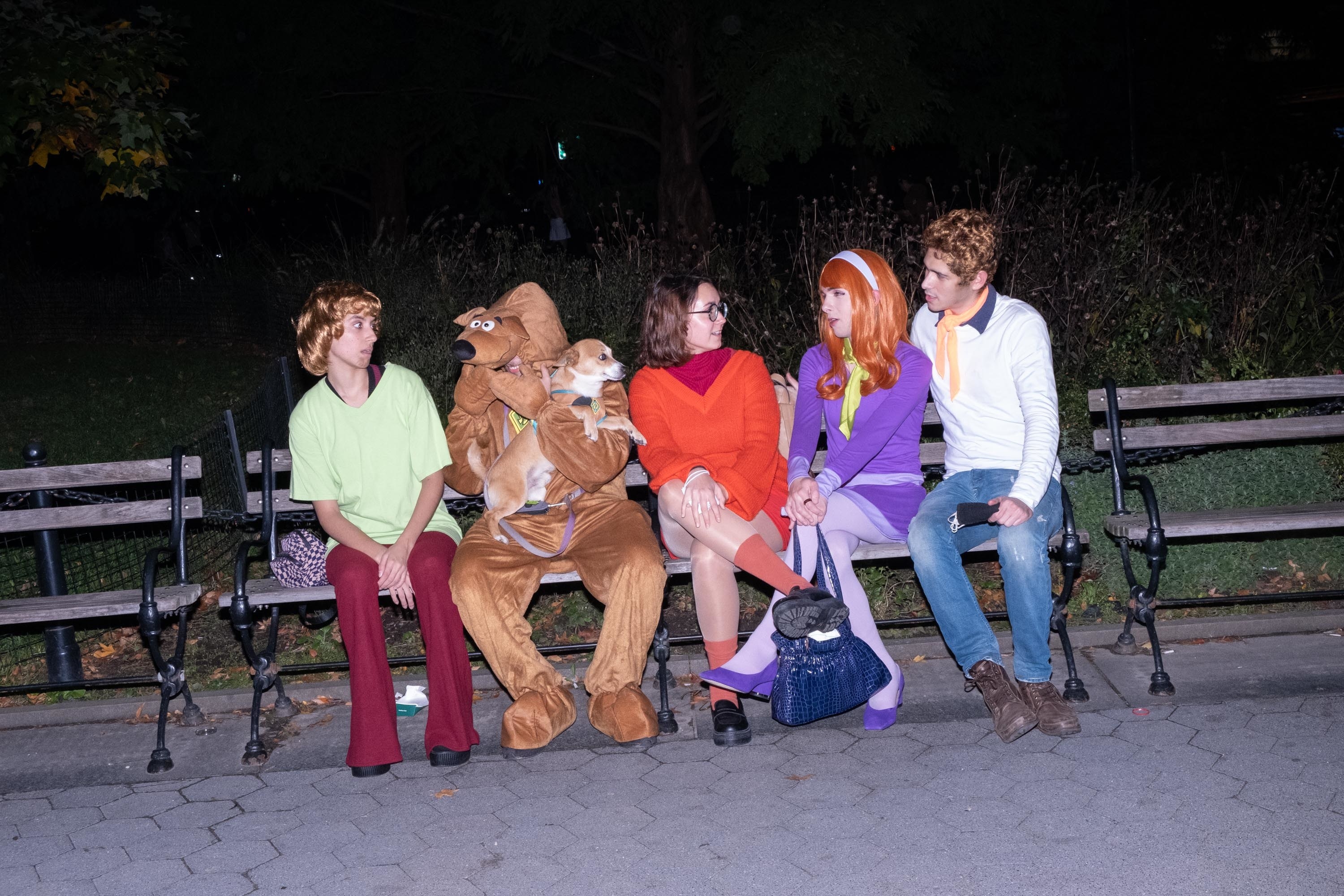 Five people dressed as the Scooby-Doo gang take a seat on a Washington Square Park bench with a small dog