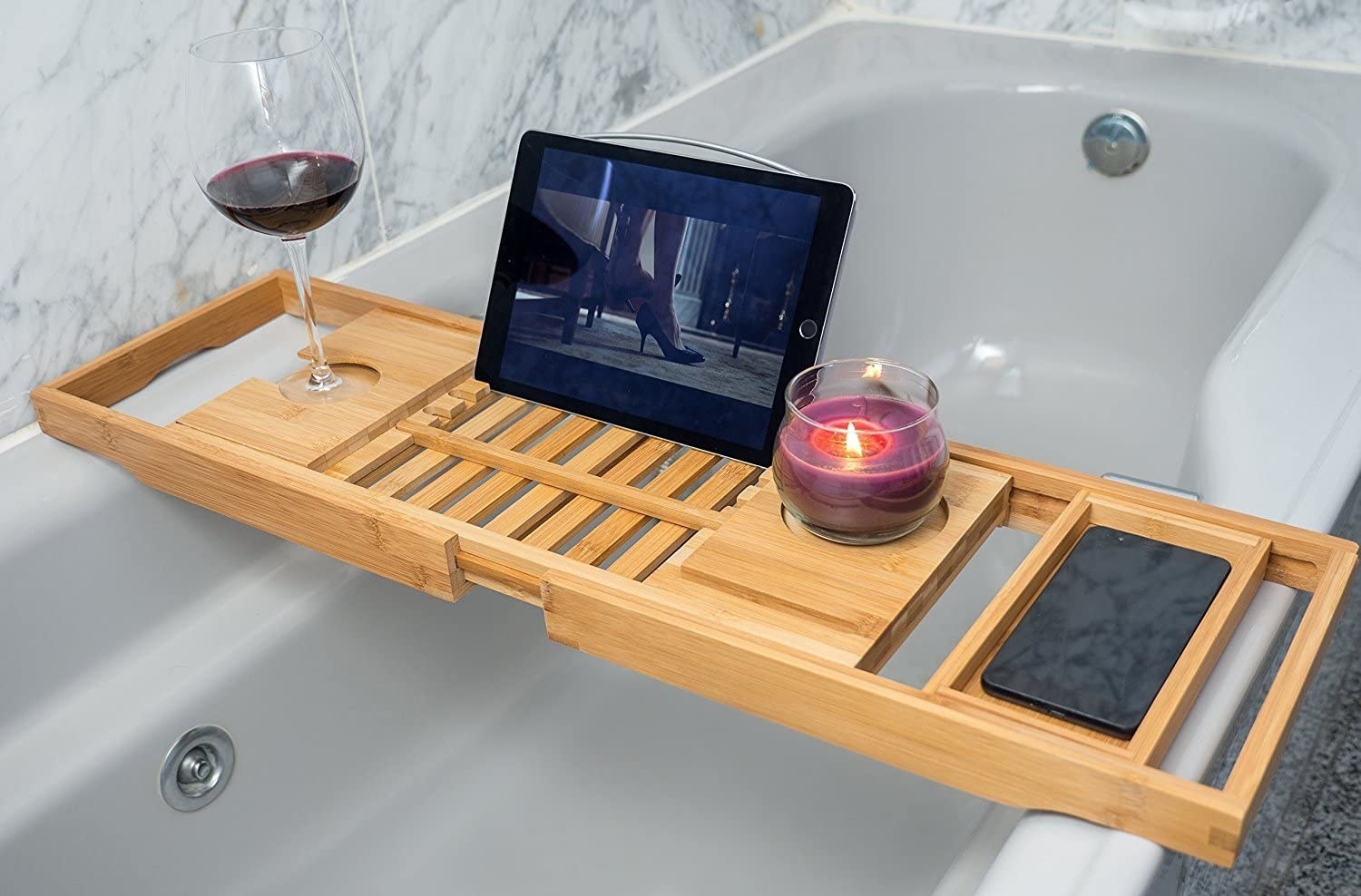 an extendable bamboo bath tray holding a tablet, wine glass, and candle