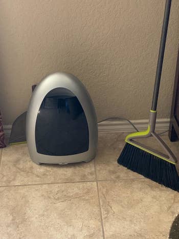silver bagless, touchless vacuum next to yellow broom on tiled floor