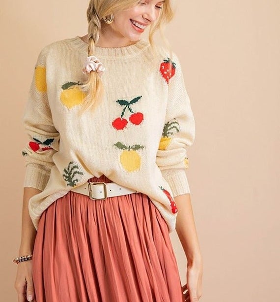 A model wearing a white sweater with colorful fruit designs, paired with a long pink skirt and white belt