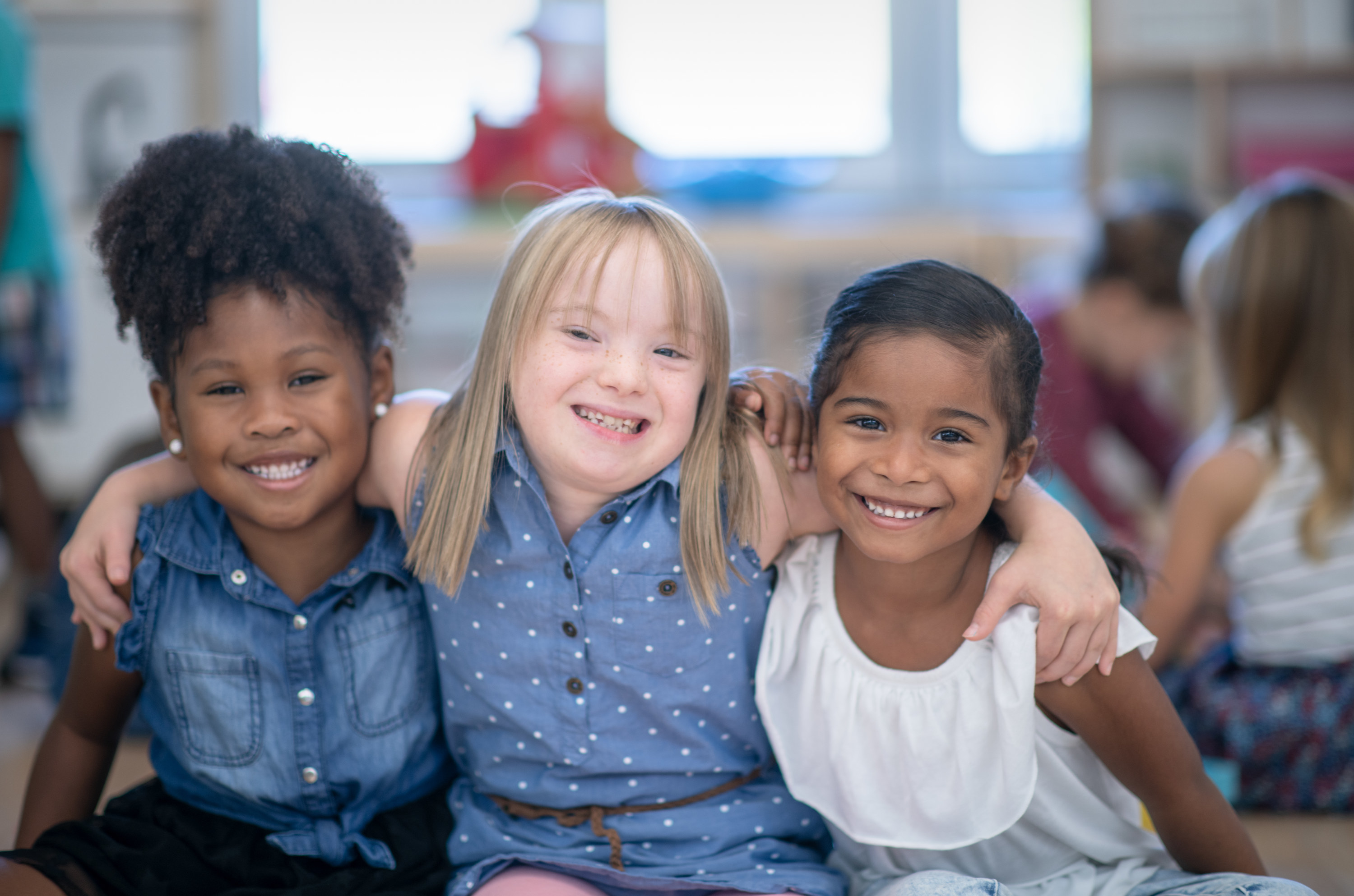 Three multi-ethnic preschool girls smiling in their classroom, and one of the girls (in the middle) has Down syndrome