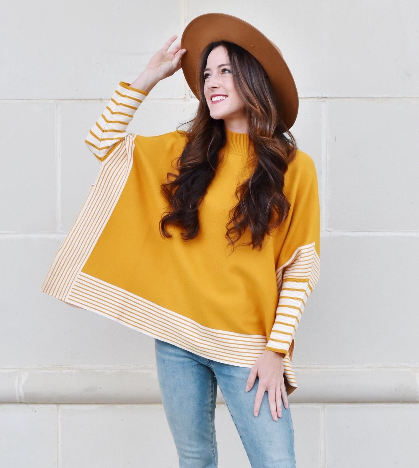 A model wearing the yellow and white poncho sweater paired with light blue jeans and a brown hat