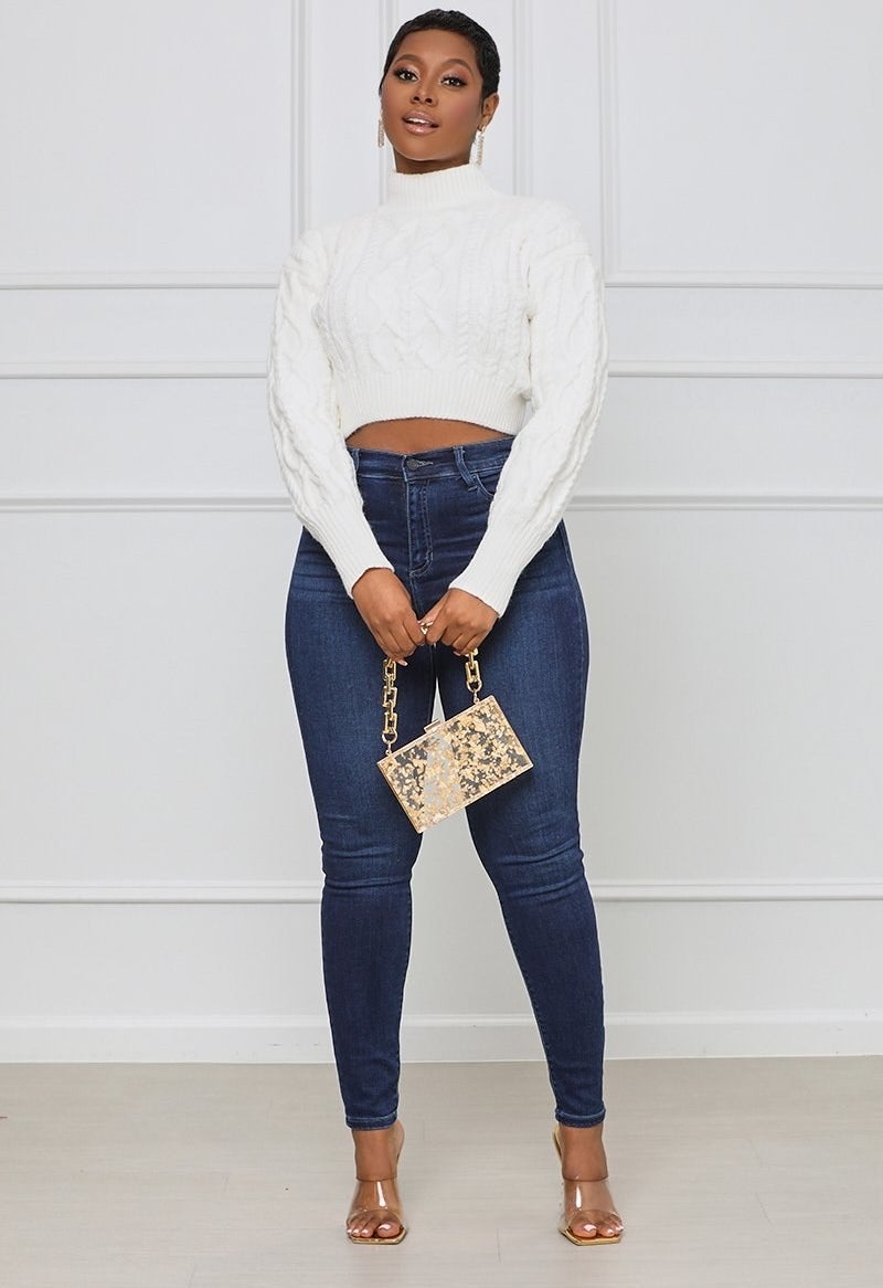 A model wearing the white cable knit crop top with high-waisted blue jeans and a gold purse