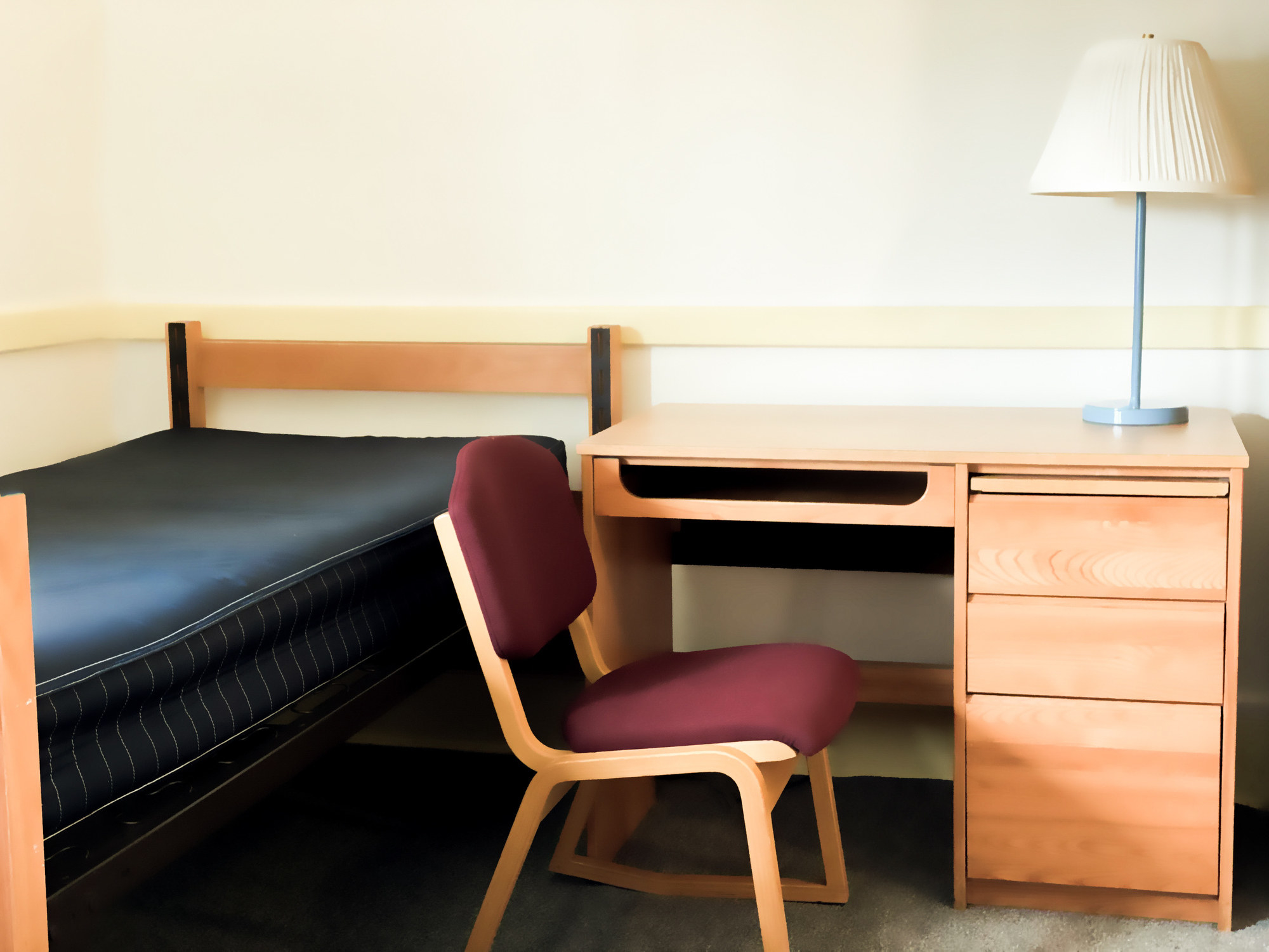 A bed sits in a student dormitory with a chair beside it near a desk with a lamp on top of the desk