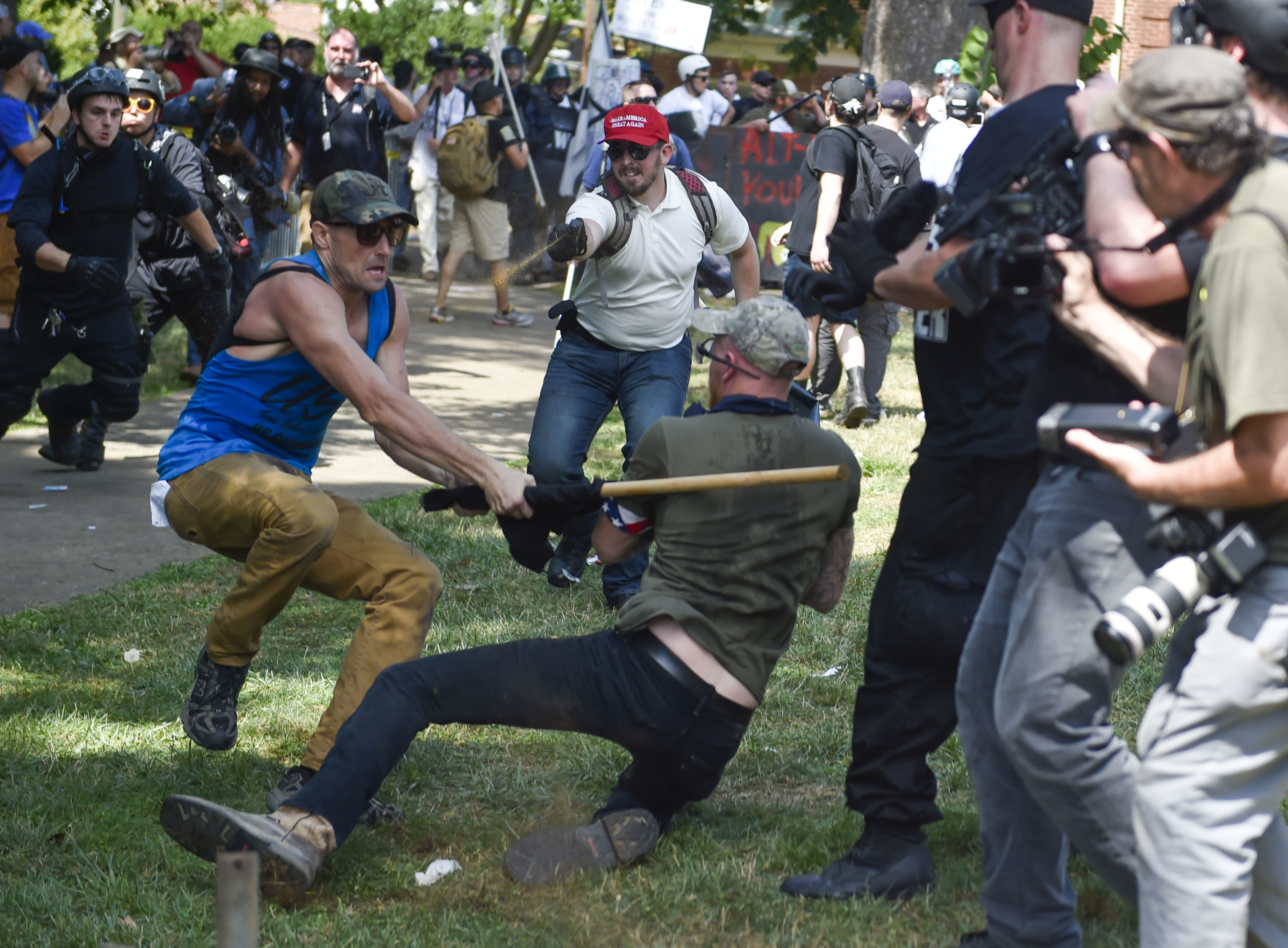 A man hits a bat into the back of another man, who is falling to the ground, while a third man wearing a Make America Great Again hat runs toward the scene and sprays the man wielding the bat