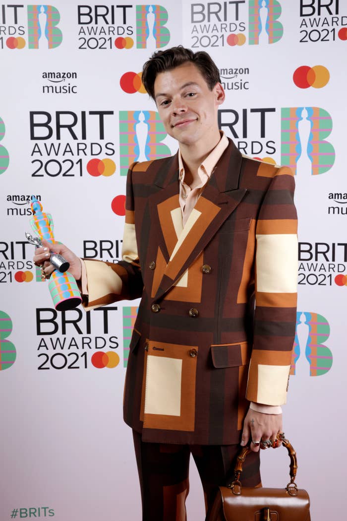 Harry at the 2021 Brit Awards holding his award as he wears a color blocked suit and carries a matching purse
