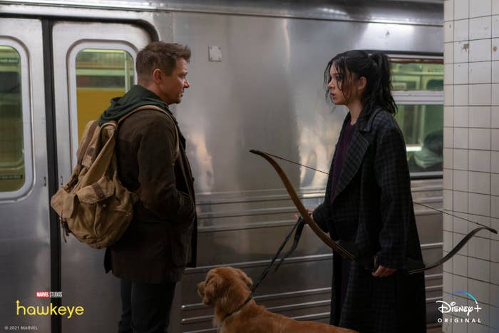 Jeremy Renner wearing backpack with Hailee Steinfeld holding a bow and a dog accompanies them.
