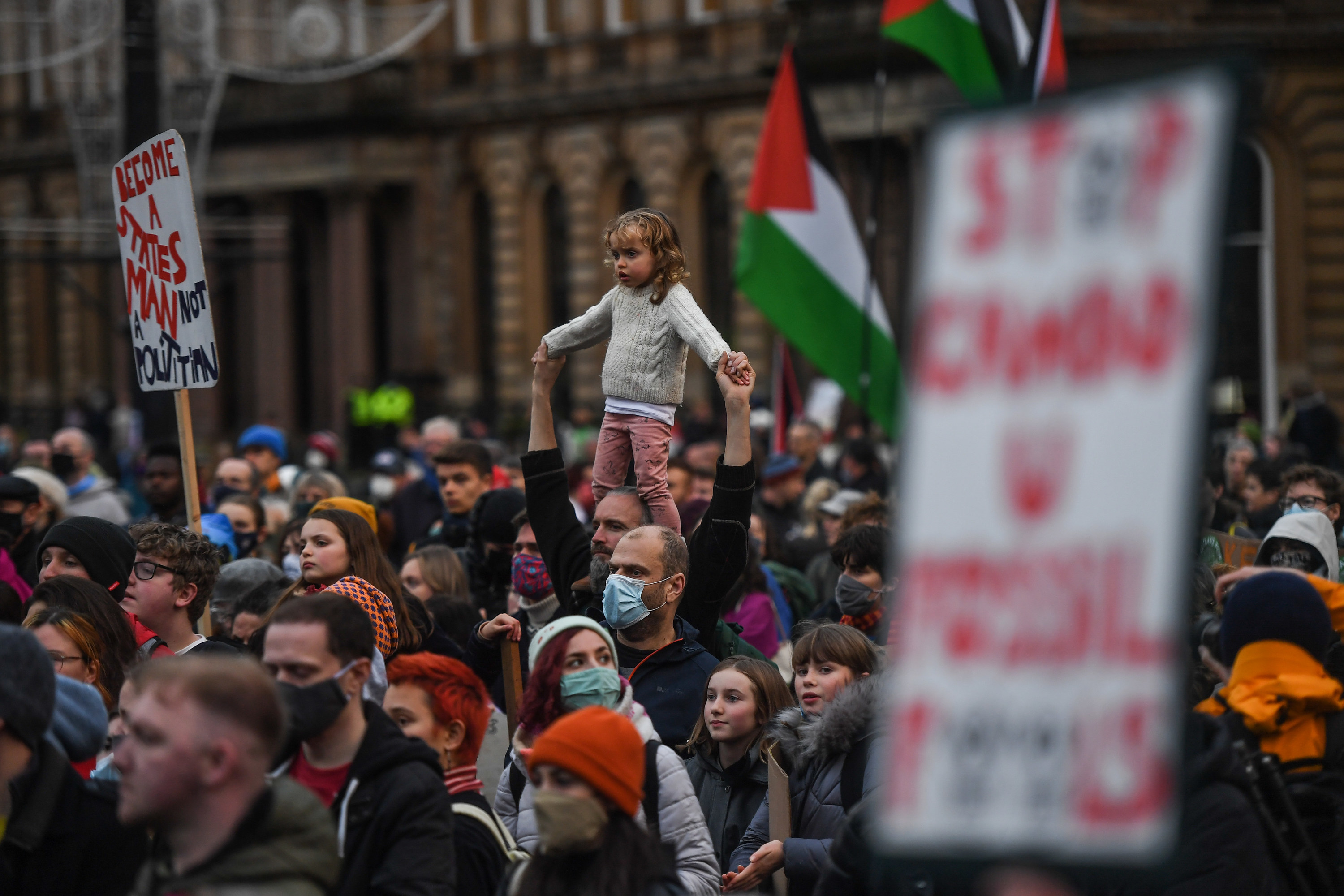 A young girl stands on an adult&#x27;s shoulders in a crowd