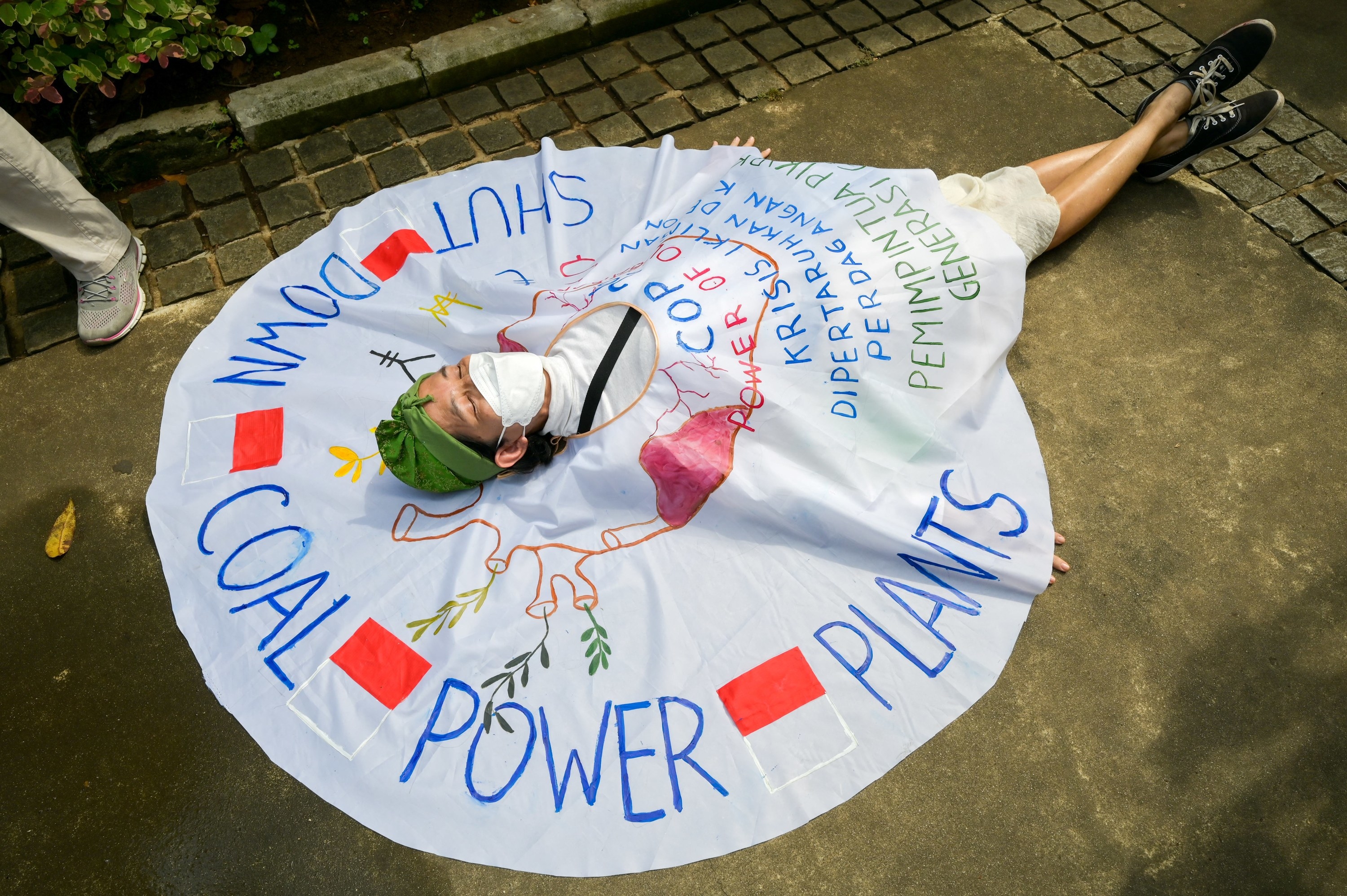 A person lies flat on the ground, their torso covered in a circular sheet that reads &quot;Shut down coal power plants&quot;