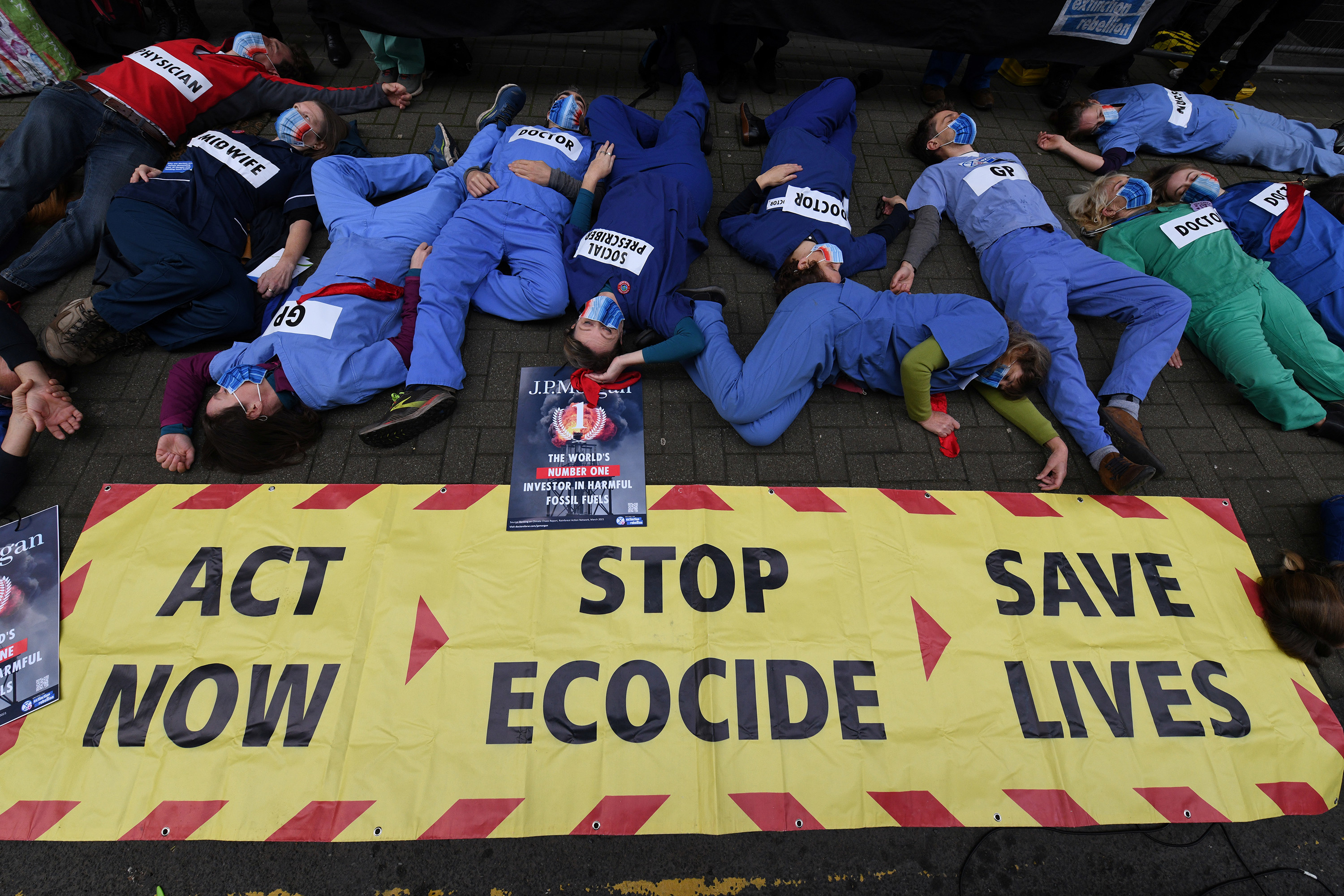 People lie on the ground next to a sign that reads &quot;Act now&quot; beside an arrow pointing to &quot;Stop ecocide&quot; beside another arrow pointing to the words &quot;Save lives&quot;