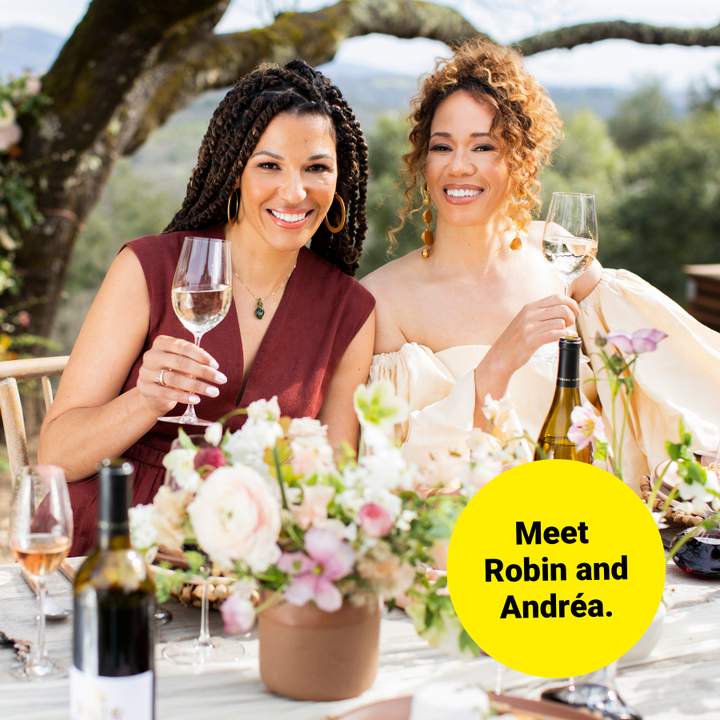 the McBride Sisters sitting at a table with flowers on it plus wine glasses and bottles. They are holding up glasses with wine in them and smiling.