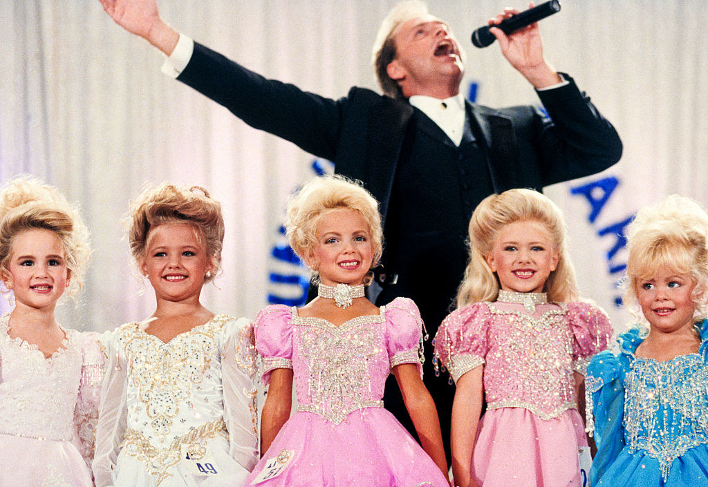 Five little blonde girls dressed in elaborate gowns and hairdos lined up as they compete in a pageant with the host standing behind them