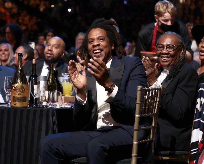Jay Z applauds while sitting in a chair next to a table with bottles of champagne on it