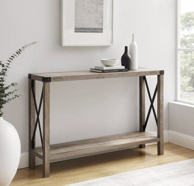 a grey-beige thin console table with black metal hardware, holding decor on its top shelf with an empty bottom shelf