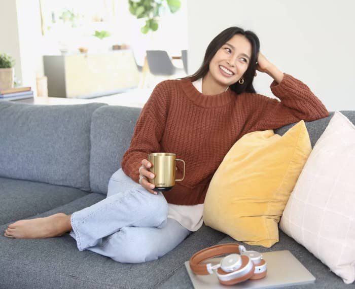 An adult sits on a couch in a sweater holding the gold mug  that says &quot;ember&quot; and smiling