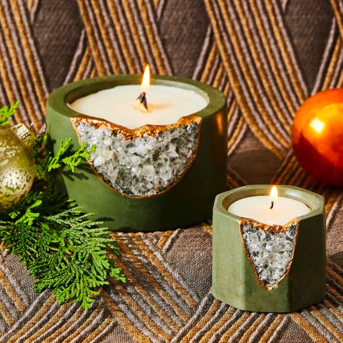 two candles in a geode vessels that have a deep evergreen shade plus gold leaf trim and crystal openings for the geode. the candles are small and large and both are lit.