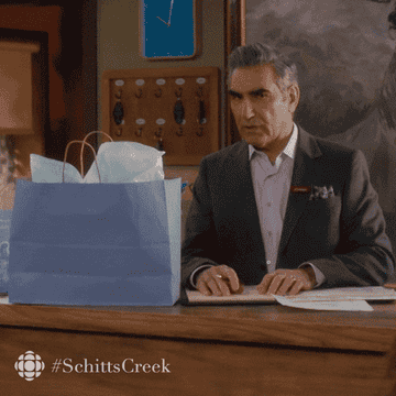 Eugene Levy peeking excitedly at a gift on the table in front of him