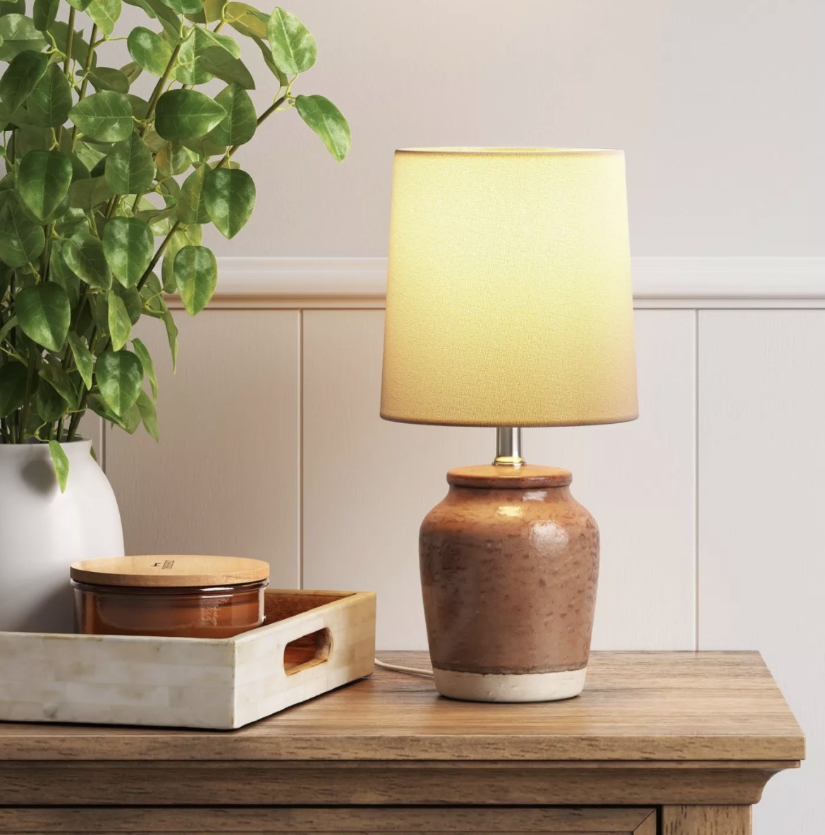 The lamp has a deep brown colored textured base with a small strip of neutral at the bottom and is turned on with a warm glow coming out of the drum shade