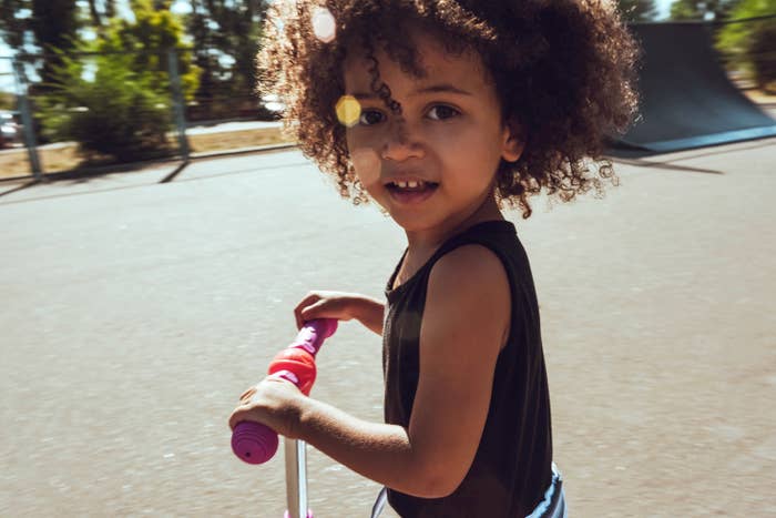Child with loose, curly hair on a scooter in a playground looks at the camera