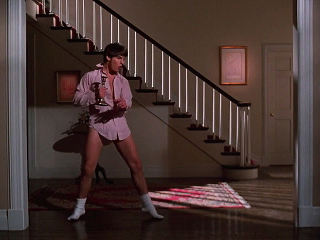 A young man dancing around his living room wearing a shirt and boxers