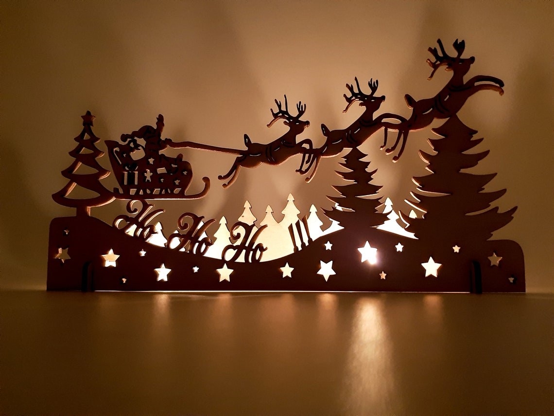 A laser-cut wood Christmas decoration depicting Santa and his reindeer flying over a forest at night, with cutouts for light from the candles to shine through