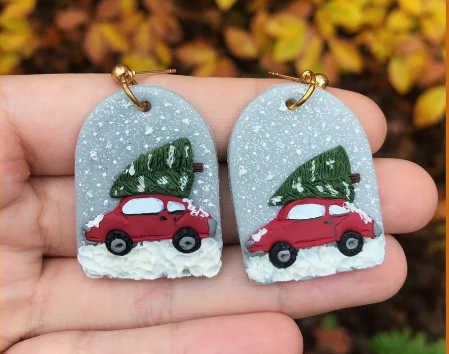 A hand holding up a pair of Christmas earrings depicting a red car with a newly chopped Christmas tree on it on a snowy day
