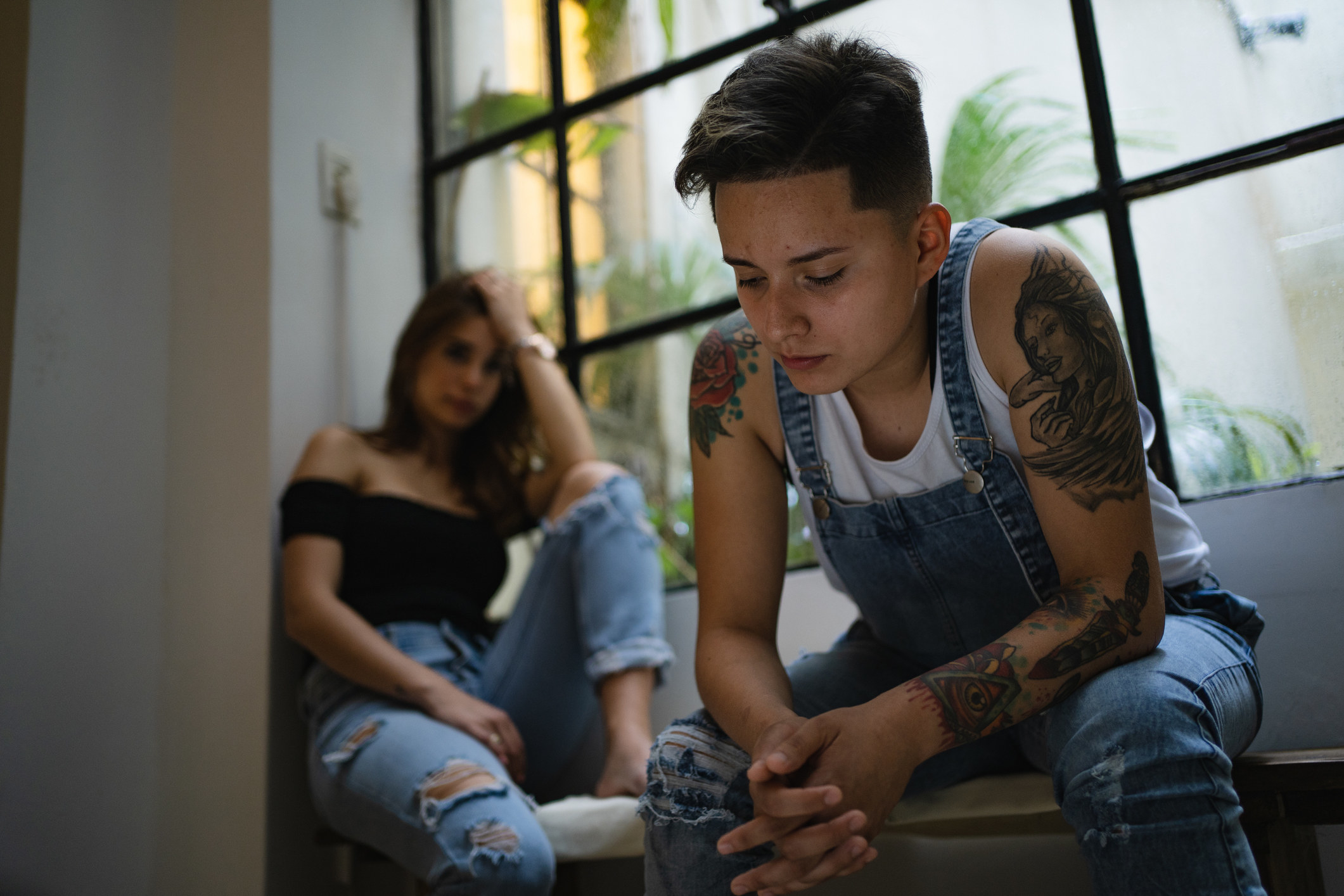 Two women sit on a couch, far apart from each other, and are in a sad state