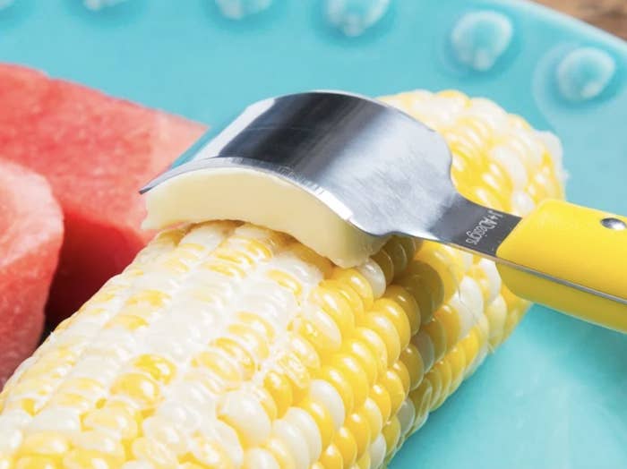 yellow and silver curved tool evenly rubs butter slice across fresh corn cob