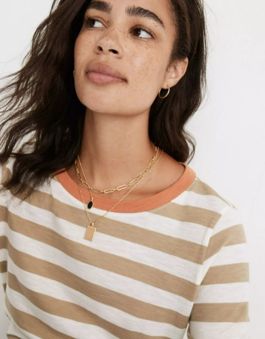 model wearing the necklace set with short-length chain necklace, medium-length black stone necklace, and long-length brass pendant necklace over a striped brown and white shirt
