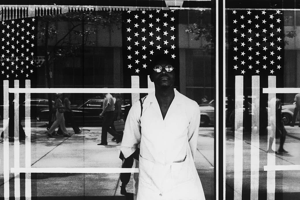 A man in sunglasses standing in front of an American flag display, with people reflected in the window behind 
