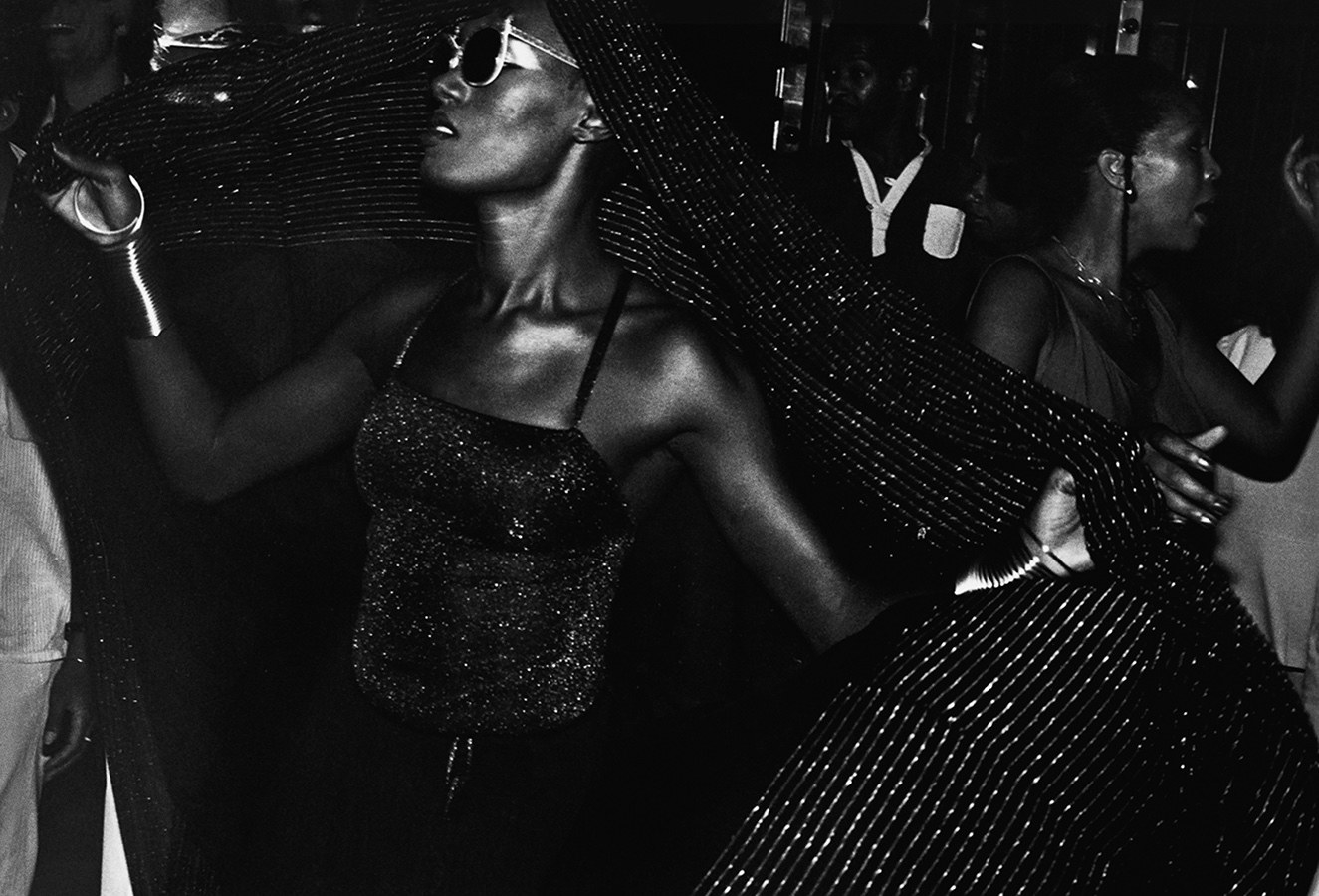 Grace Jones in a sparkly top and wrap dancing with sunglasses, people in the background