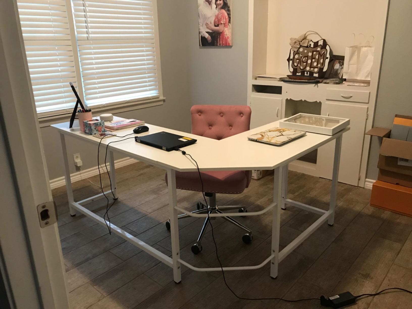 White L-shaped desk in front of pink office chair with laptop and office supplies spread out