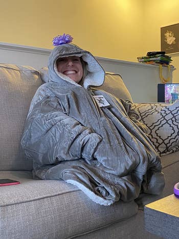 A reviewer wearing the blanket sweater in gray sitting on the couch with a bow on their head