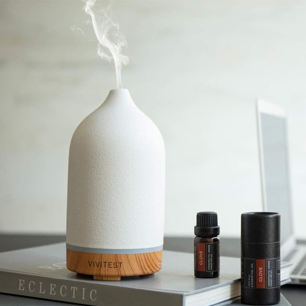 The 100ml diffuser sitting on a book beside two essential oil bottles. The diffuser has a wooden base and a cream-colored top.