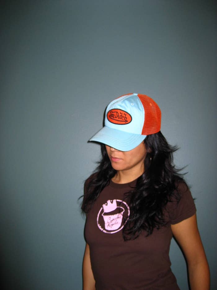 A woman with long brown hair wears a baby blue and dark orange Von Dutch trucker cap, which obscures her eyes.