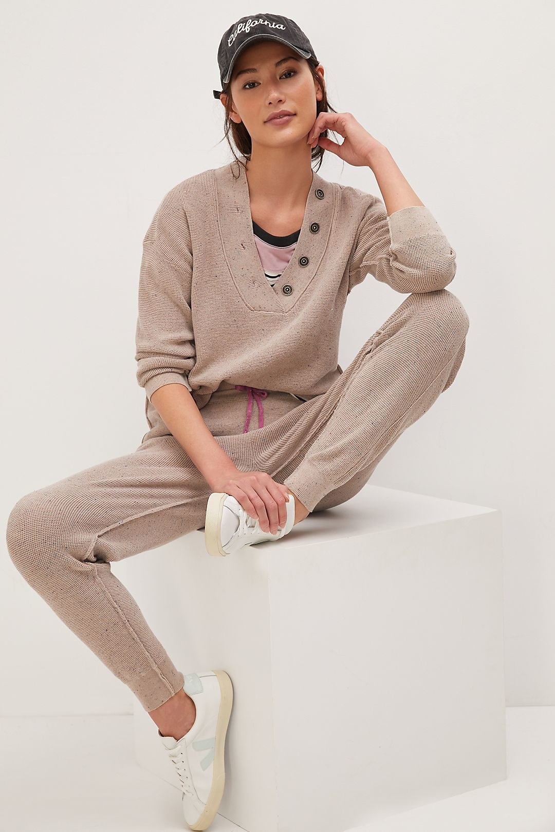 model wearing oatmeal colored knit loungewear set, and baseball had and sneakers