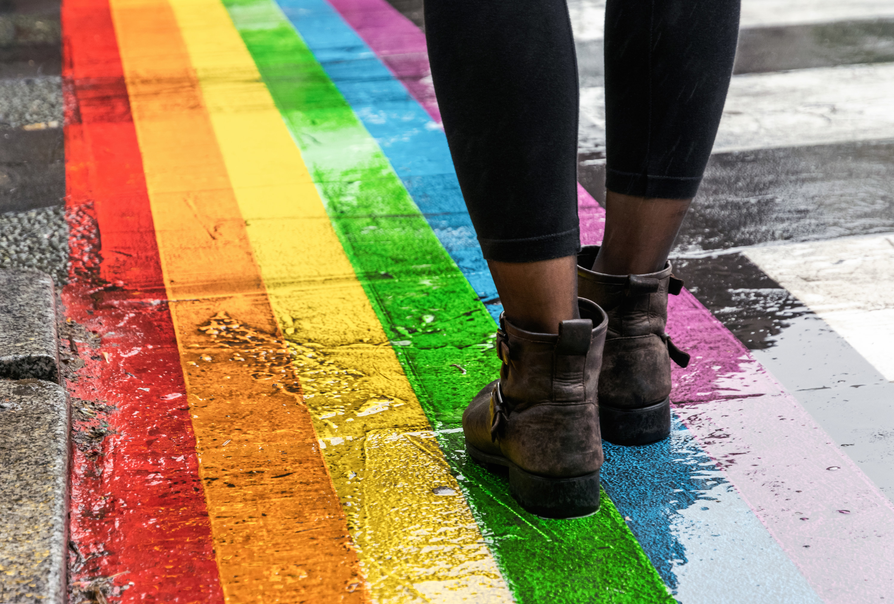 Brown boots and black leggings standing on a rainbow painted crosswalk in the rain.