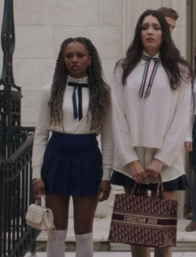 Monet Hann wears a long sleeve shirt tucked into a dark skirt and knee length socks. Luna La wears an oversized knit sweater over a button up shirt with a dark skirt