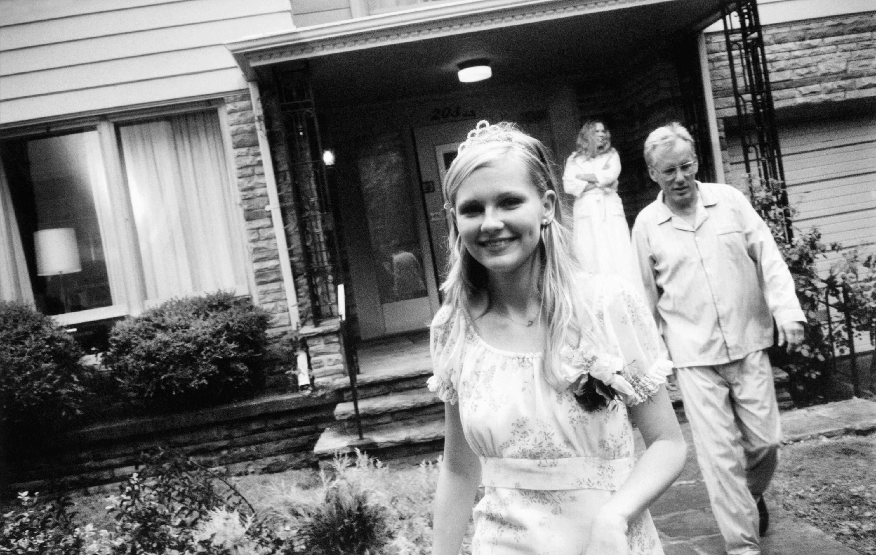 Dunst wears a dress and tiara while walking in front of a house with James Woods behind her