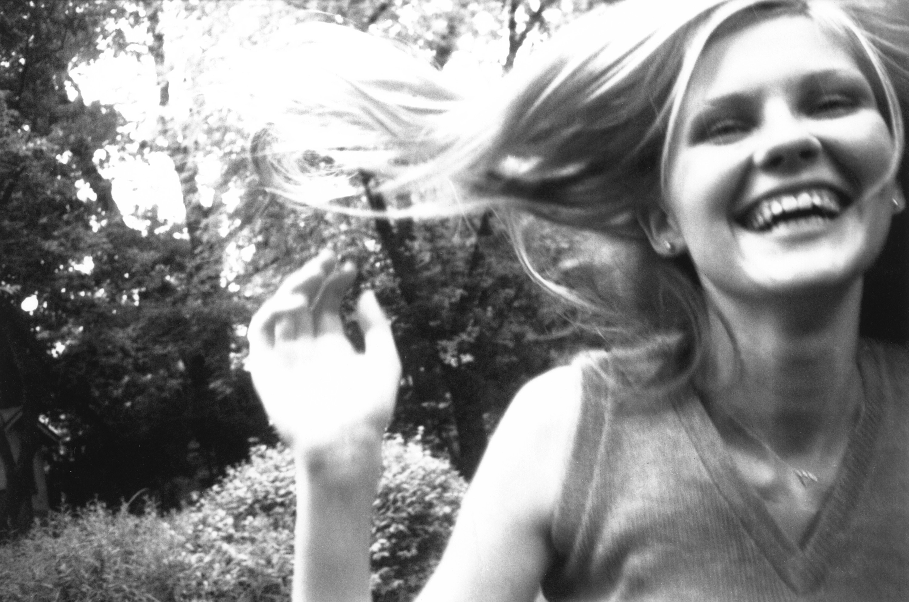 Dunst smiles with her hand up and her hair blowing in the wind with trees in the background