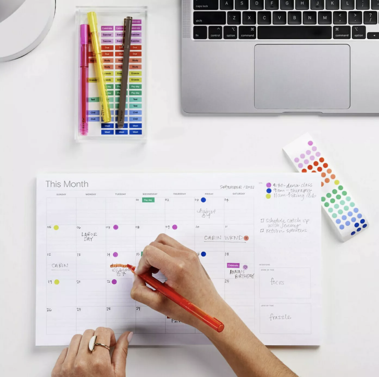 A person makes color-coded notes on a desktop monthly calendar.