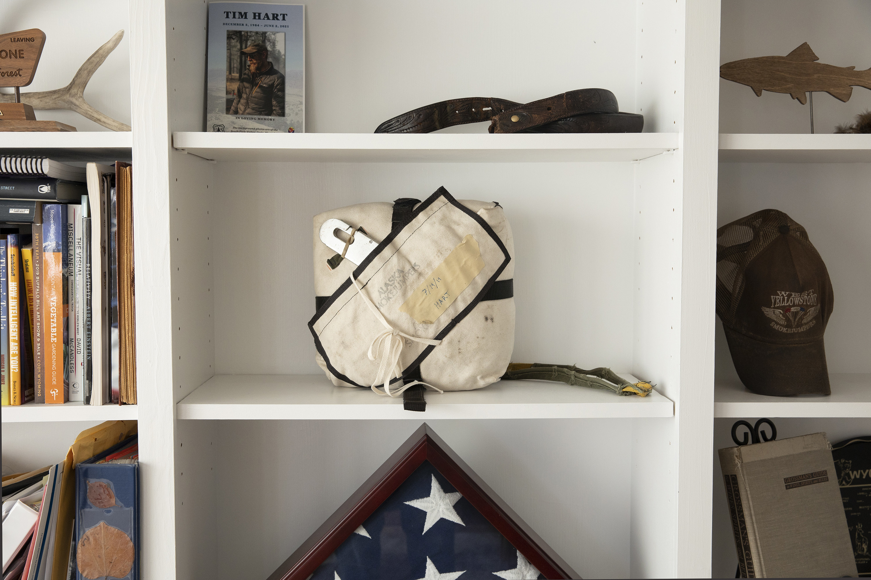 A bookcase holds a pamphlet for tim hart, his parachute, and an american flag