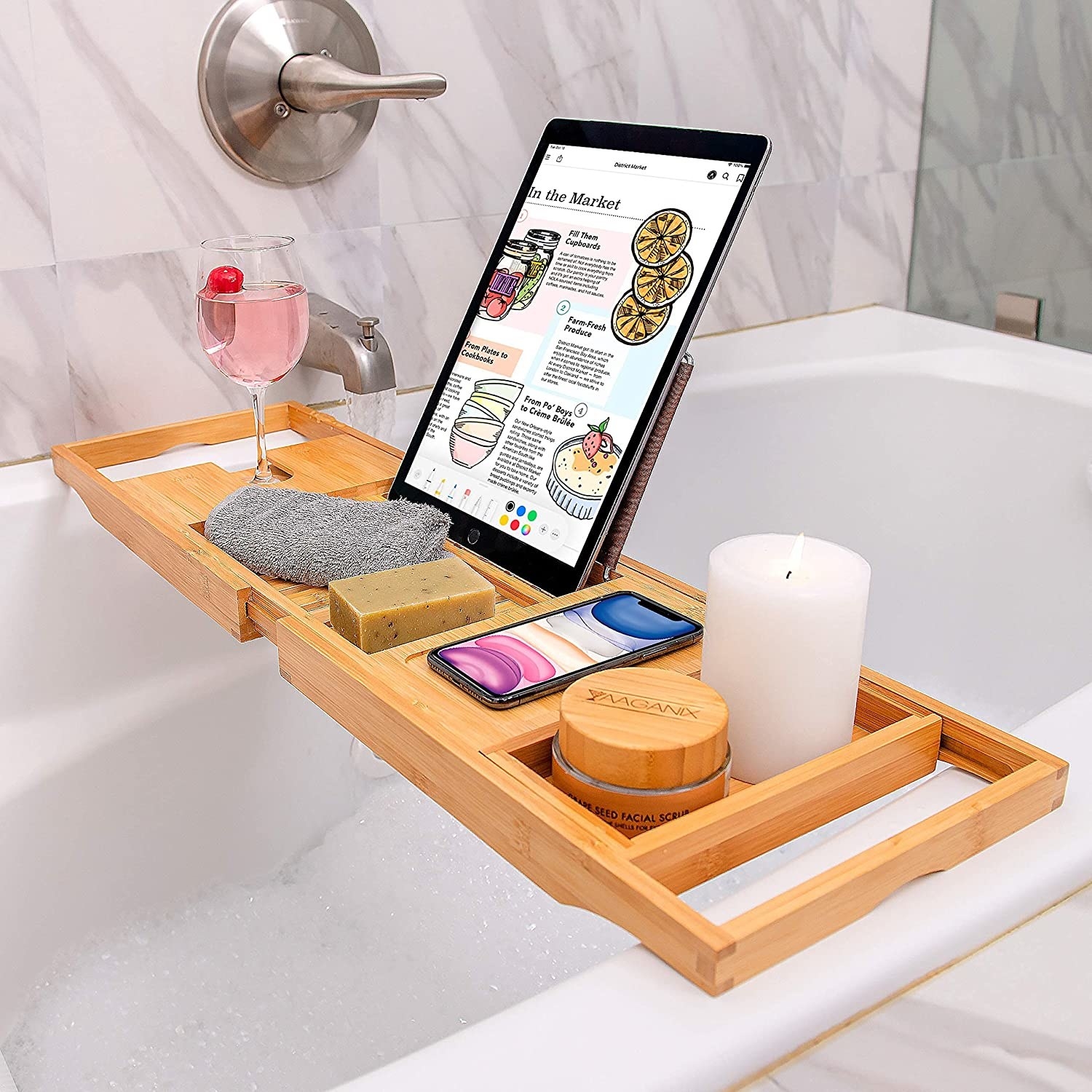 The wooden bath tray holding a tablet, wine glass, phone, candle, soap, face scrub, and small towel