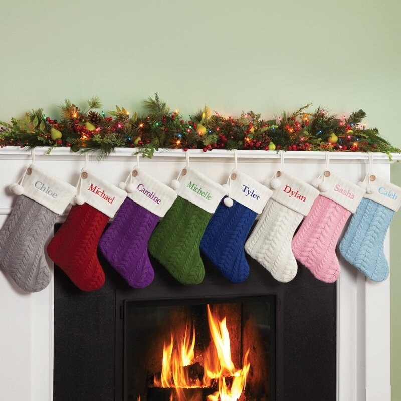 from left to right: gray, red, purple, green, blue, white, pink, light blue personalized stockings hanging above mantle, fireplace lit