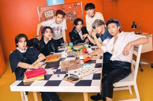 BTS poses in a room while sat at a table covered in board games and books