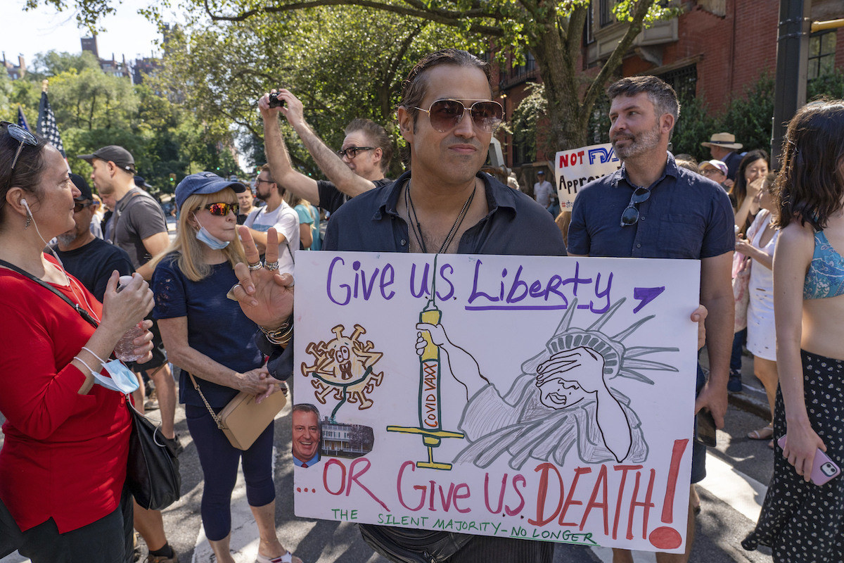 A man holds a sign that reads &quot;give us liberty or give us death&quot; with an illustration of the statue of liberty holding a COVID vaccine syringe in disgust