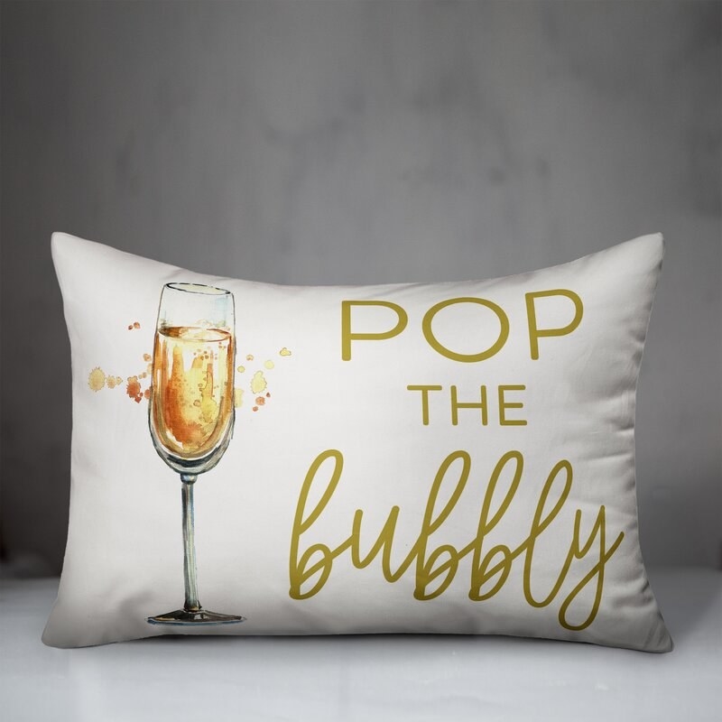 White pillow with champagne glass on it and &quot;pop the bubbly&quot; in gold lettering