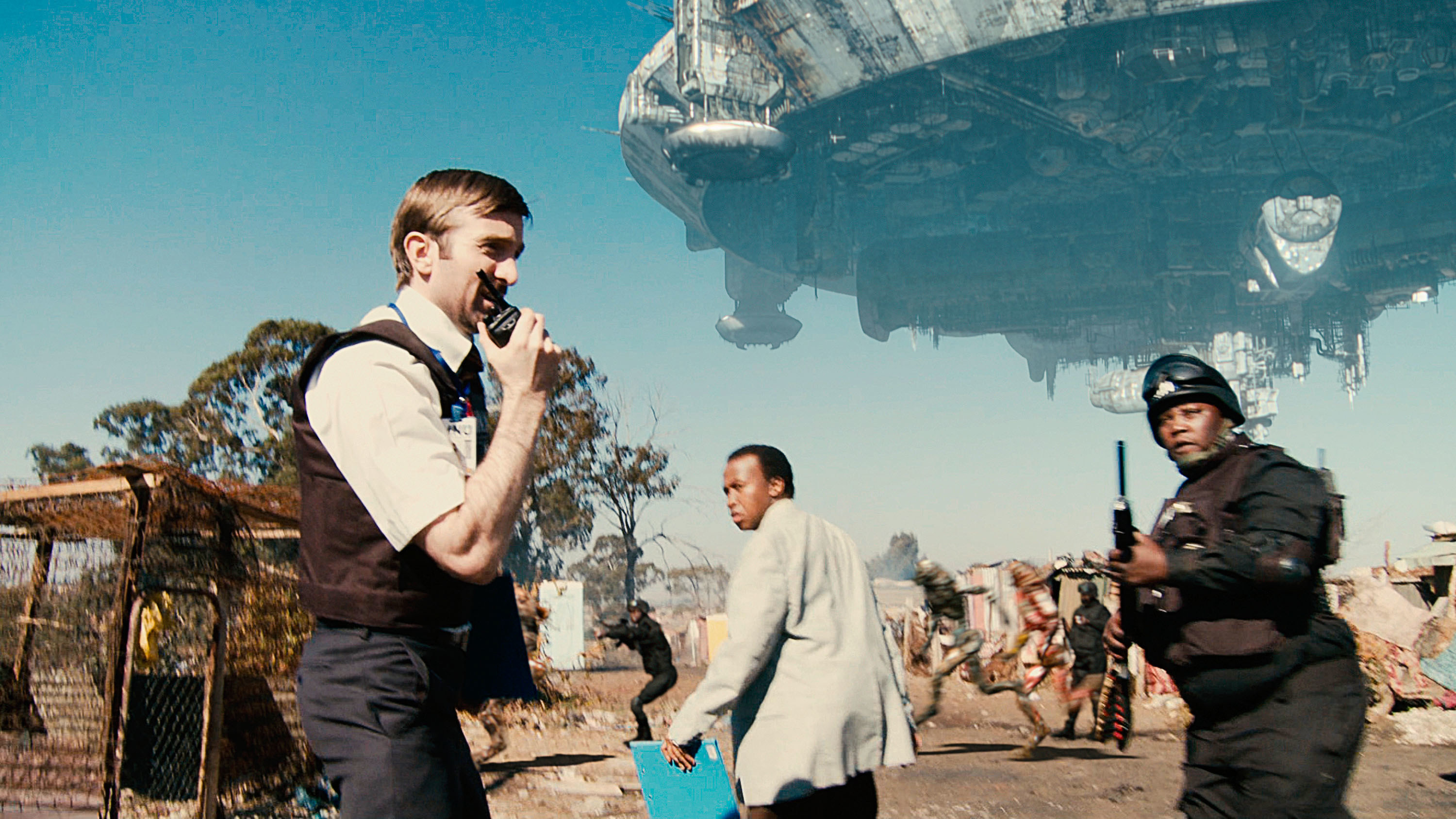 Sharlto Copley, Mandla Gaduka, Kenneth Nkosi dressed as police officers, a giant alien ship in the sky above them